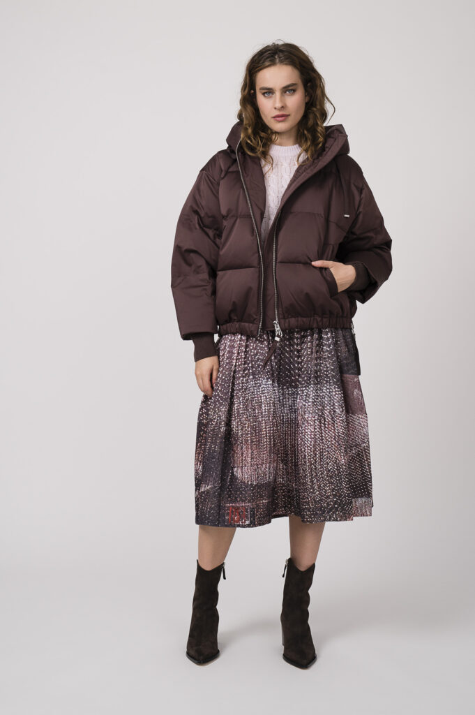 Model zeigt eine bordeaux-rote Daunenjacke von DUNO, einen Kurzarmpullover in rose der Marke Dorothee Schumacher und einen bordeaux-rot gemusterten Rock der Marke Talking Walls.