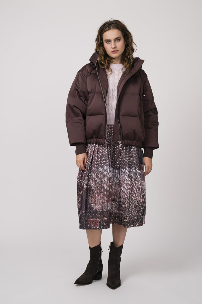 Model zeigt eine bordeaux-rote Daunenjacke von DUNO, einen Kurzarmpullover in rose der Marke Dorothee Schumacher und einen bordeaux-rot gemusterten Rock der Marke Talking Walls.