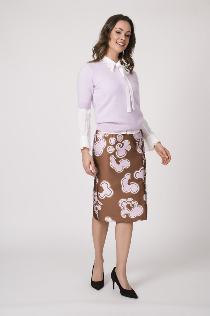 Model trägt eine weiße Bluse, eine Flieder farbigen Kurzarmpullover und einen beige rosa gemusterten Bleistiftrock der Marke Dorothee Schumacher.