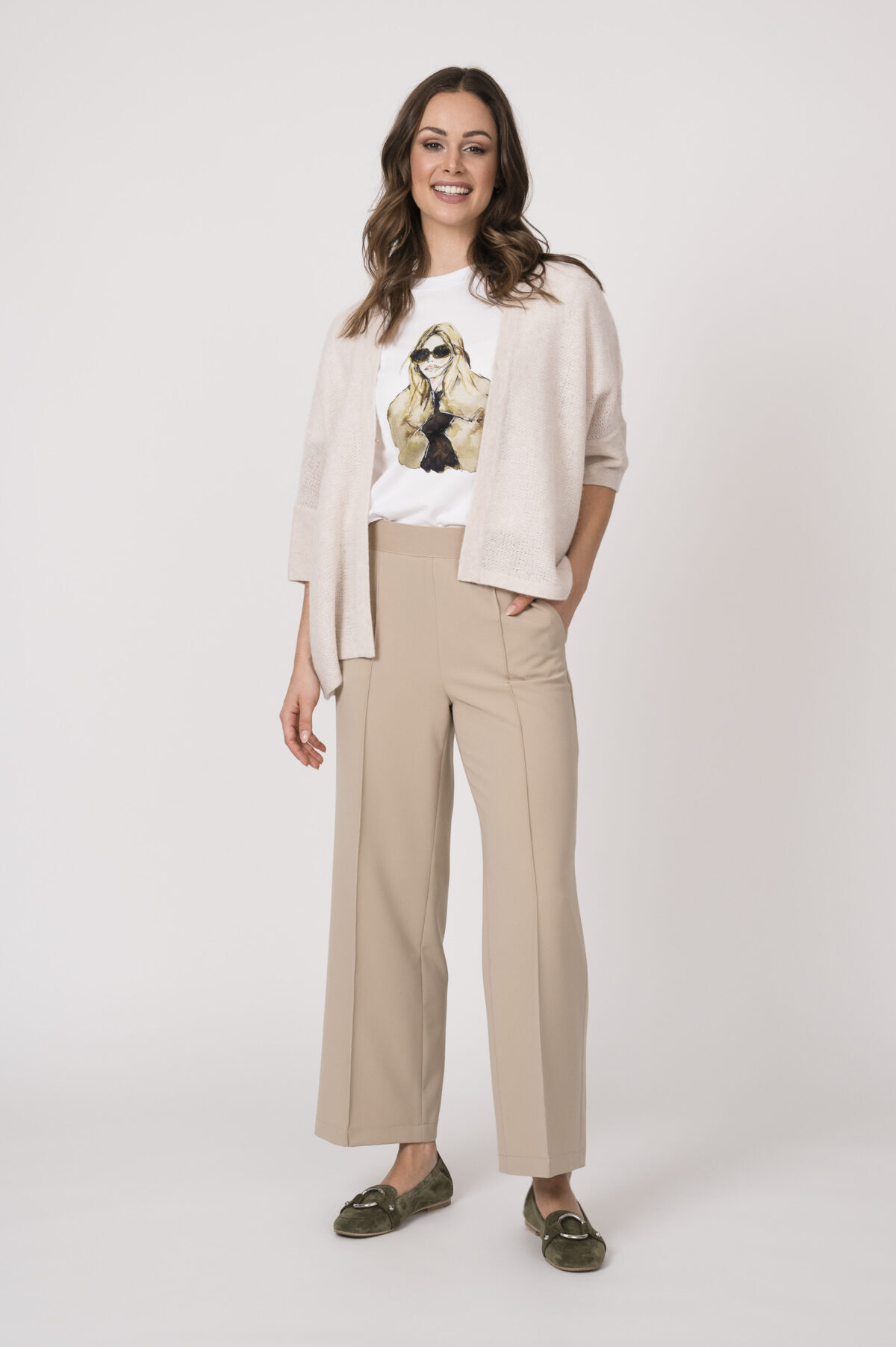 Model trägt eine sand-farbige Strickweste der Marke Hemisphere, ein weißes Shirt mit Zeichnung einer Frau der Marke Unartig und eine beige Hose der Marke Cambio.