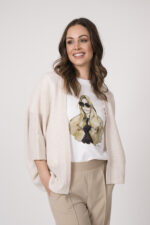 Model trägt eine sand-farbige Strickweste der Marke Hemisphere, ein weißes Shirt mit Zeichnung einer Frau der Marke Unartig und eine beige Hose der Marke Cambio.