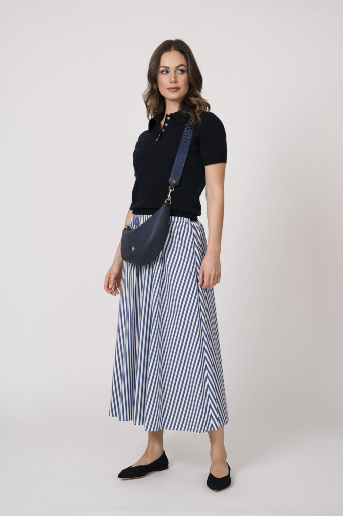 Model trägt einen marine-blauen Kurzarmpullover der Marke Allude und einen blau weiß gestreiften Rock der Marke JOOP sowie einer dunkelblauen Halbmondtasche der Marke Aigner.
