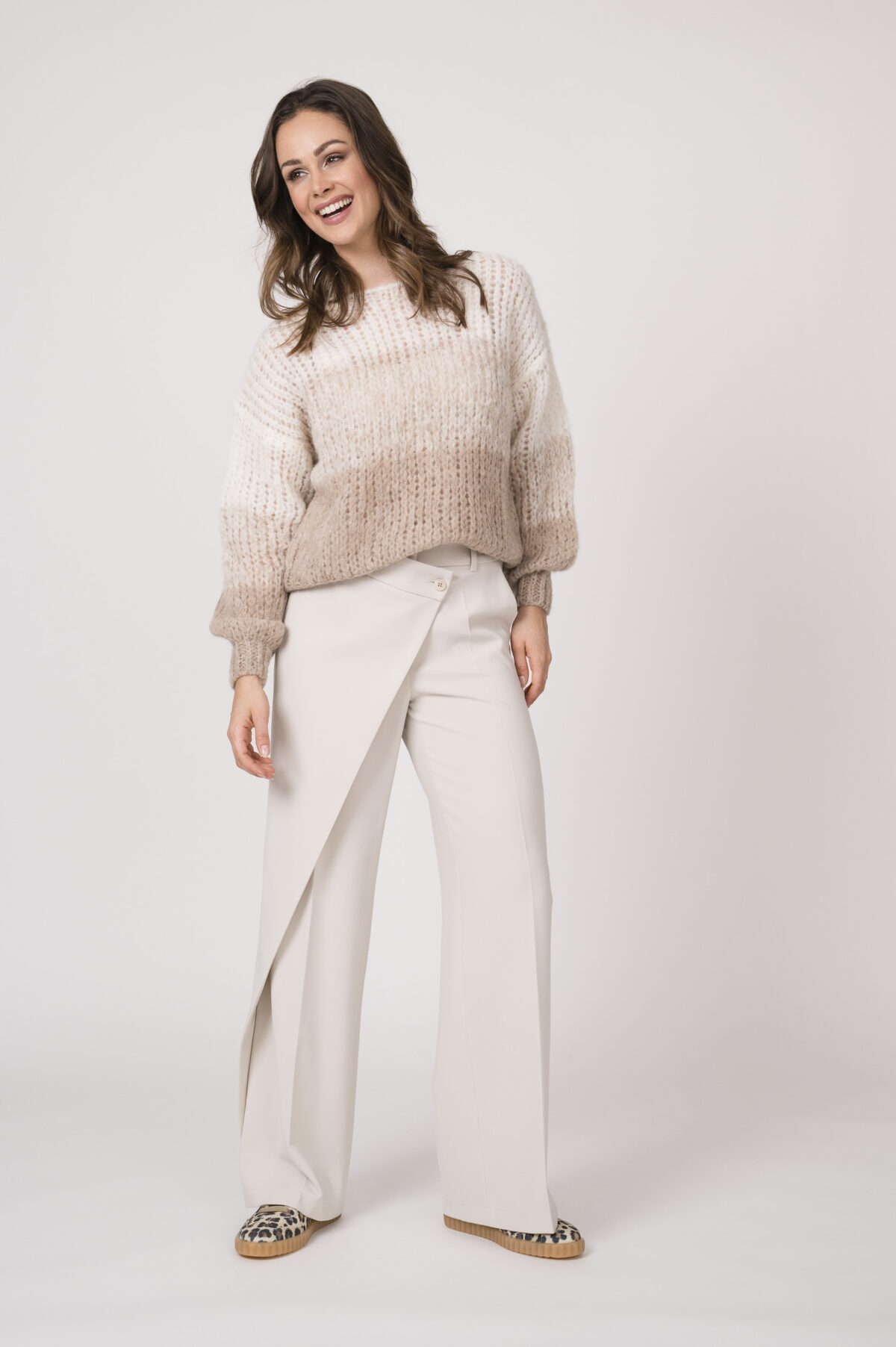 Model trägt einen hellen Strickpullover der Marke Michelle & Hoven und eine natur-farbige Stoffhose der Marke Cambio sowie Sneaker in Leo-Print der Marke Voile Blanche.