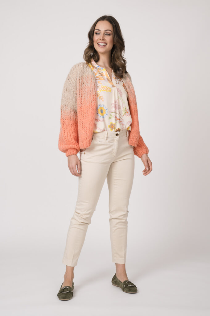 Model trägt eine beige-gemusterte Bluse der Marke Herzens und eine helle Strickweste der Marke Michelle & Hoven sowie eine sand-farbige Stoffhose der Marke Mason’s.