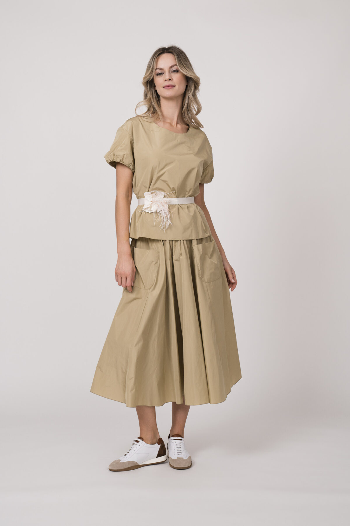 Model trägt ein beiges Outfit der Marke Susanne Bommer bestehend aus: Kurzarm-Oberteil, Rock und Federgürtel in weiß.