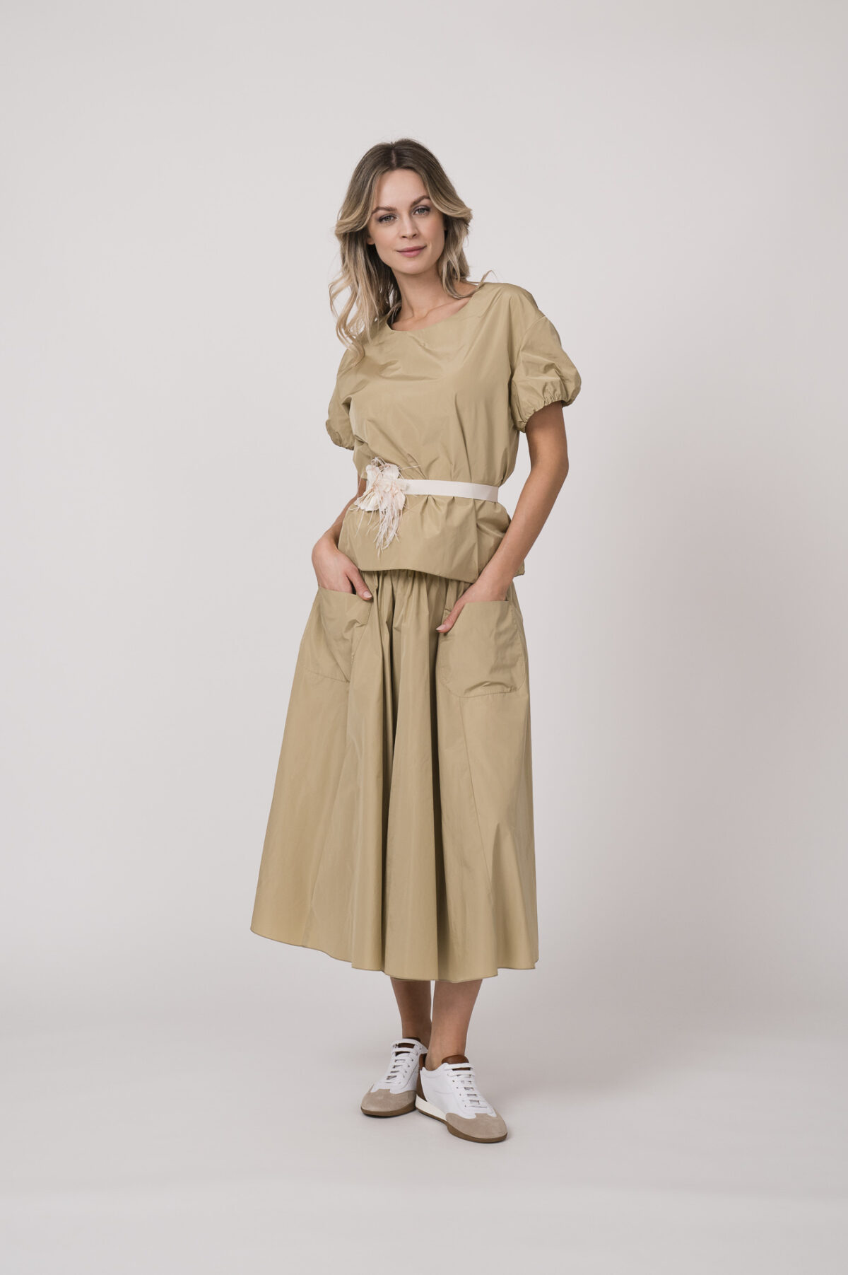 Model trägt ein beiges Outfit der Marke Susanne Bommer bestehend aus: Kurzarm-Oberteil, Rock und Federgürtel in weiß.