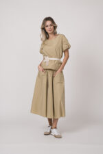 Model trägt ein beiges Outfit der Marke Susanne Bommer bestehend aus: Kurzarm-Oberteil, Rock und Federgürtel in weiß.