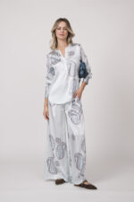 Model trägt ein Ensemble aus fließender Bluse und Hose in hellem Blau mit Paisley Muster der Marke Herzens sowie eine kleine dunkelblaue Tasche der Marke Tissa Fontaneda.