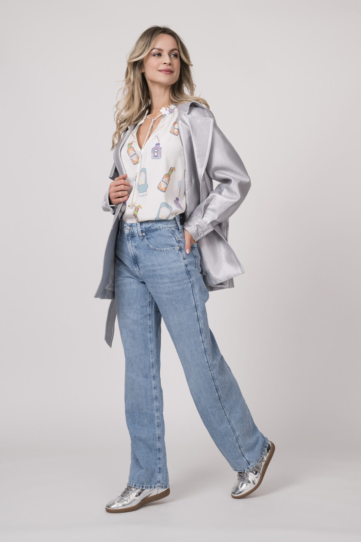 Weiße Bluse mit Flaschen-Muster der Marke Herzens, hellblaue Jeans der Marke 7 for all Mankind, kurzer silber Trenchcoat der Marke Marc Cain.