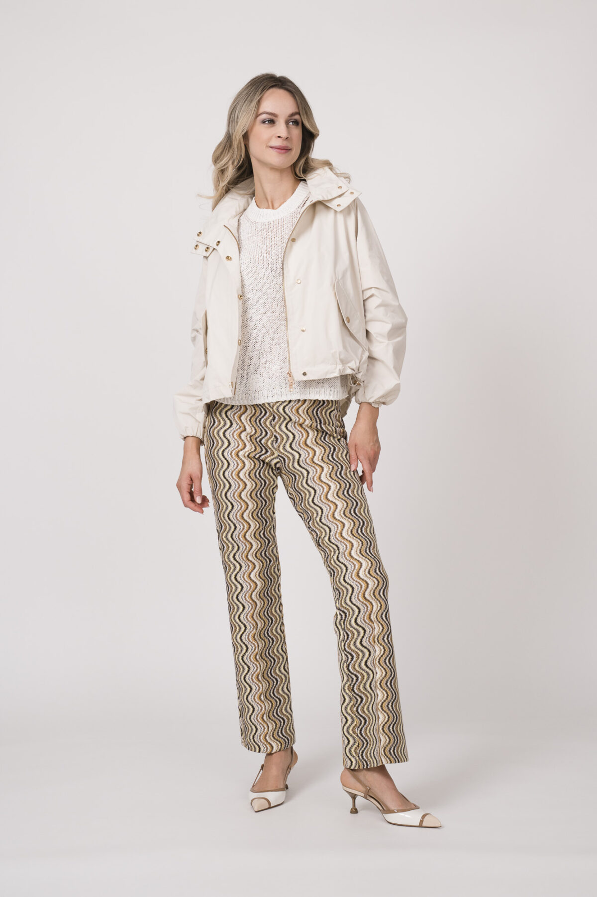 Model trägt eine Jacke der Marke Mabrun in Natur mit goldfarbenen Druckknöpfen. Dazu trägt sie eine gemusterte Hose der Marke Cambio und einen Kurzarmpullover der Marke FFC in Weiß.