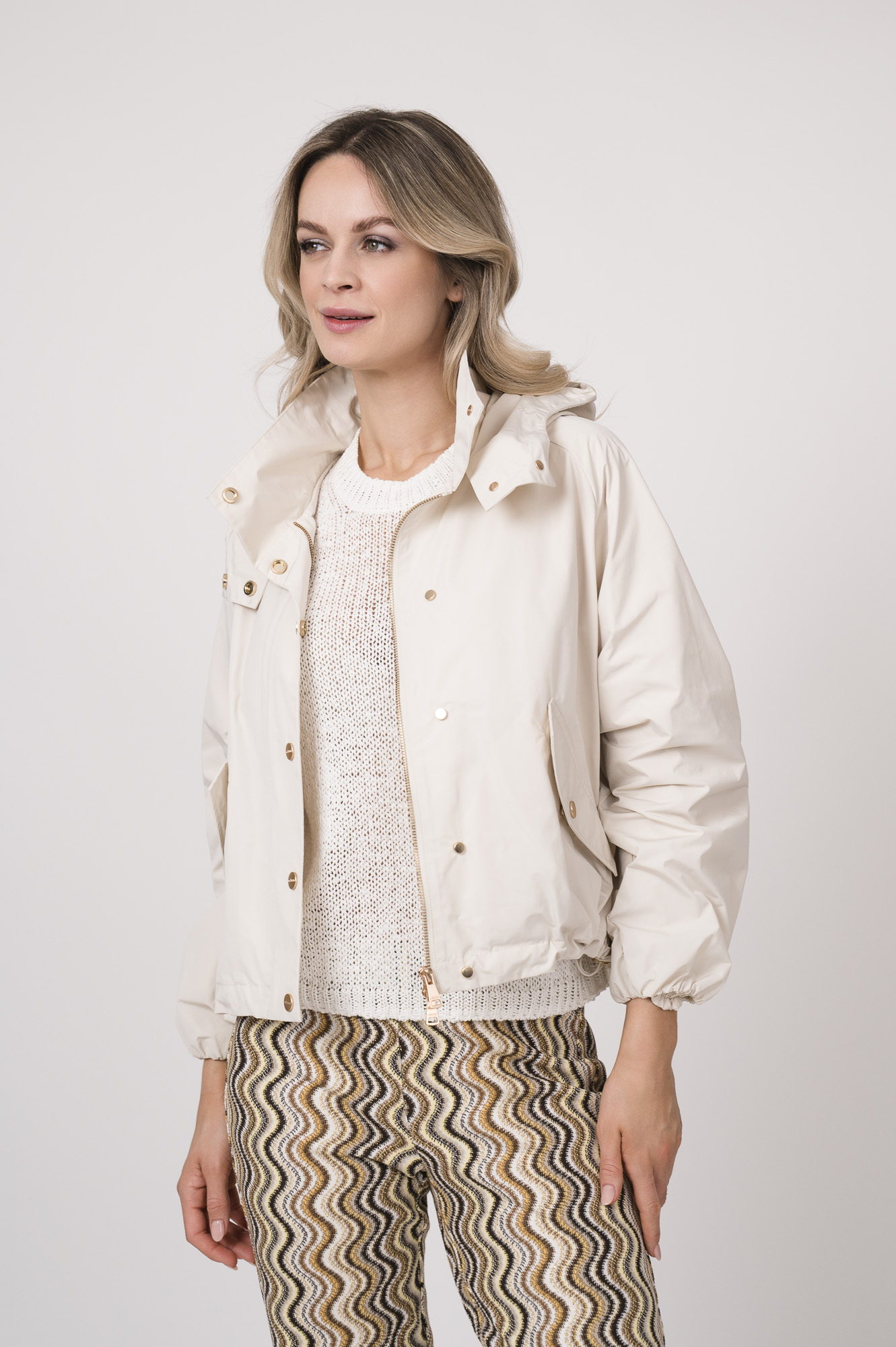 Model trägt eine Jacke der Marke Mabrun in Natur mit goldfarbenen Druckknöpfen. Dazu trägt sie eine gemusterte Hose der Marke Cambio und einen Kurzarmpullover der Marke FFC in Weiß.