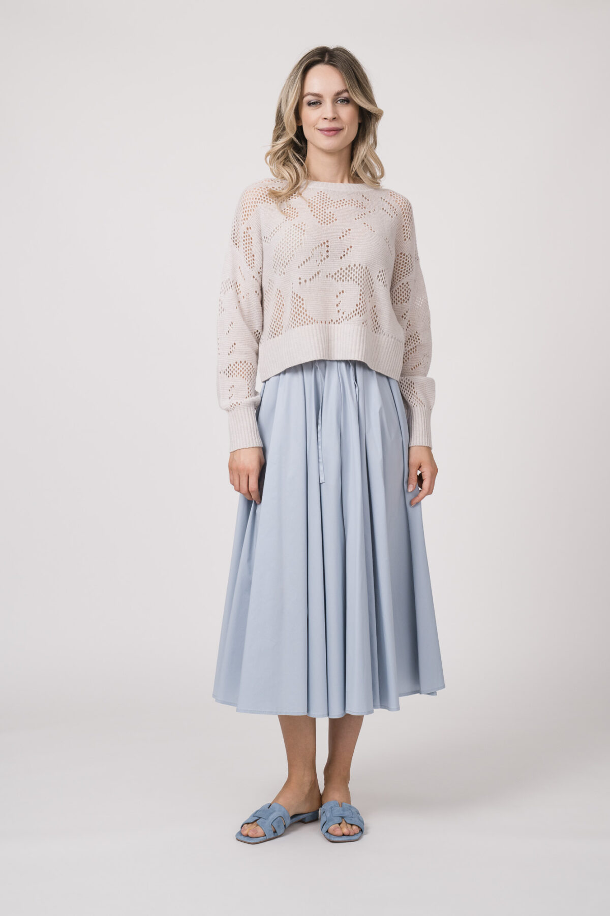 Model trägt einen Langarmpullover in der Farbe Sand der Marke Herzens, dazu einen Rock der Marke FFC in Hellblau.