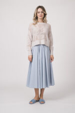 Model trägt einen Langarmpullover in der Farbe Sand der Marke Herzens, dazu einen Rock der Marke FFC in Hellblau.