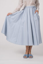 Model trägt einen Langarmpullover in der Farbe Sand der Marke Herzens, dazu einen Rock der Marke FFC in Hellblau.