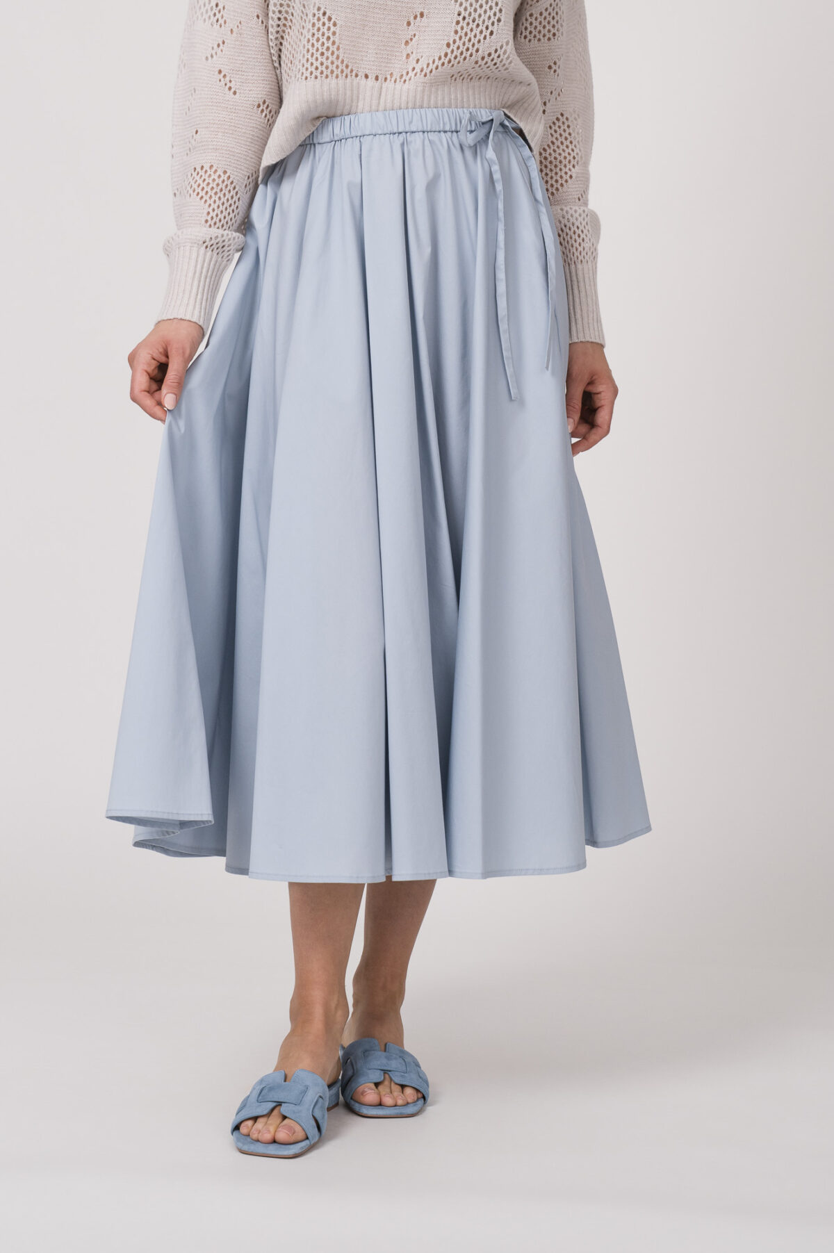 Model trägt einen Langarmpullover in der Farbe Sand der Marke Herzens, dazu einen Rock der Marke FFC in Hellblau.