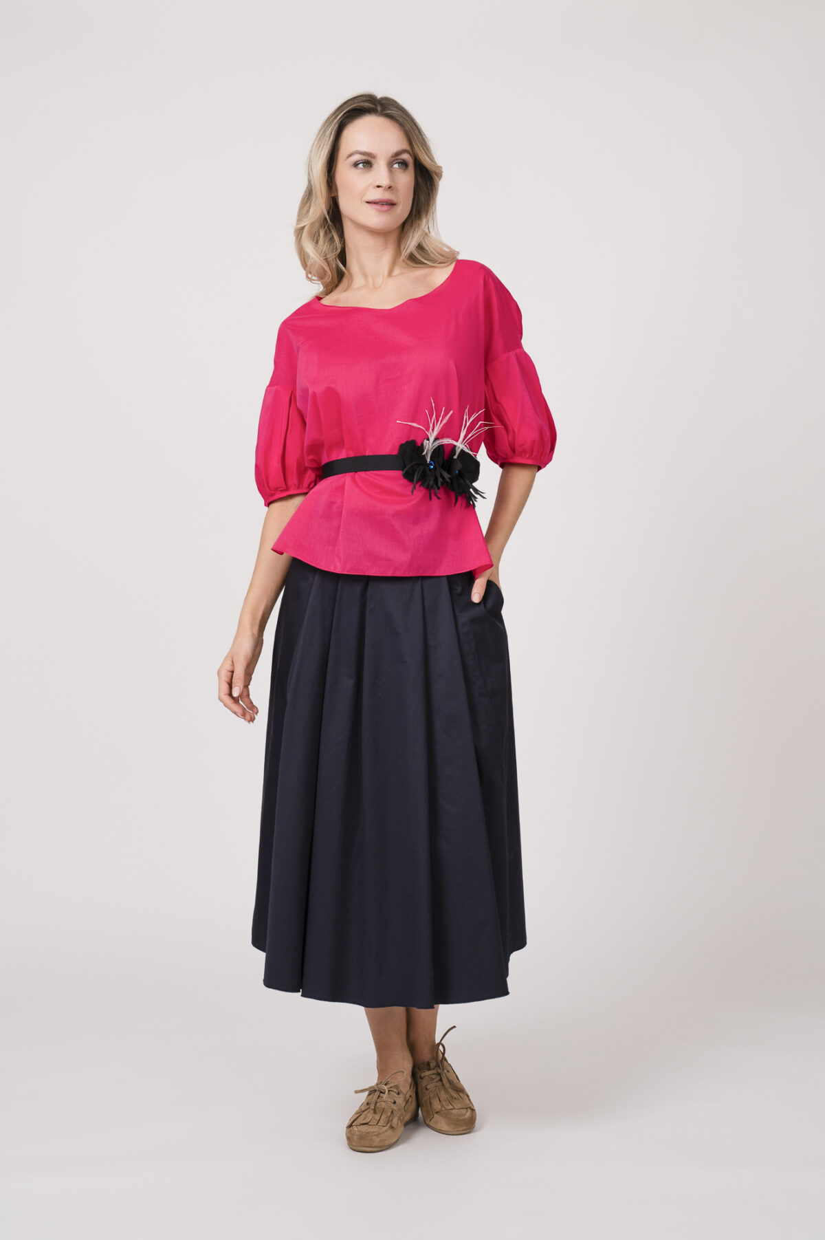 Model trägt eine Bluse der Marke Susanne Bommer in der Farbe Pink, dazu einen Rock der Marke Susanne Bommer in Marine. Einen Gürtel mit Federn der Marke Susanne Bommer.