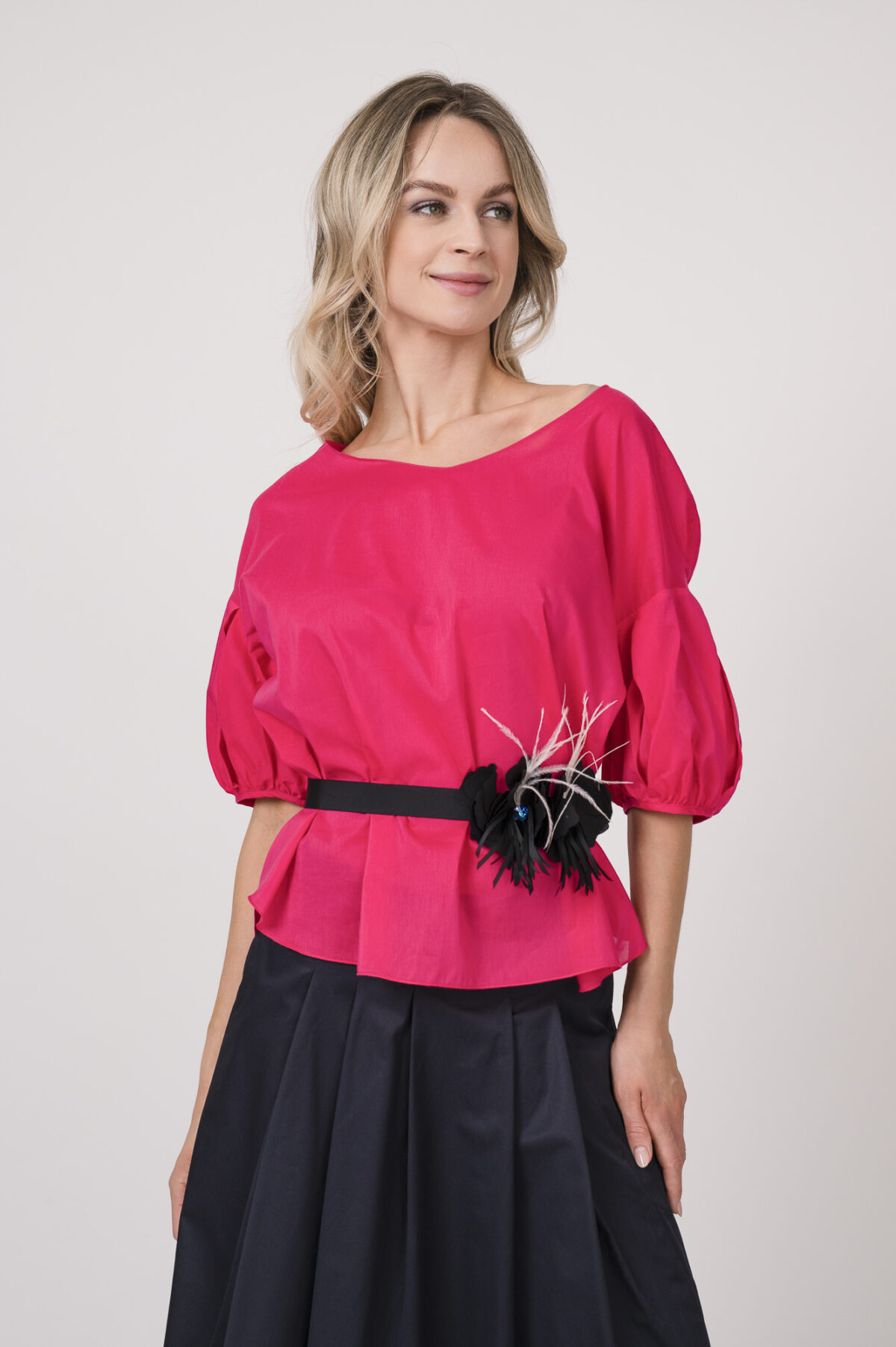 Model trägt eine Bluse der Marke Susanne Bommer in der Farbe Pink, dazu einen Rock der Marke Susanne Bommer in Marine. Einen Gürtel mit Federn der Marke Susanne Bommer.