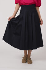 Model trägt eine Bluse der Marke Susanne Bommer in der Farbe Pink, dazu einen Rock der Marke Susanne Bommer in Marine. Einen Gürtel mit Federn der Marke Susanne Bommer.