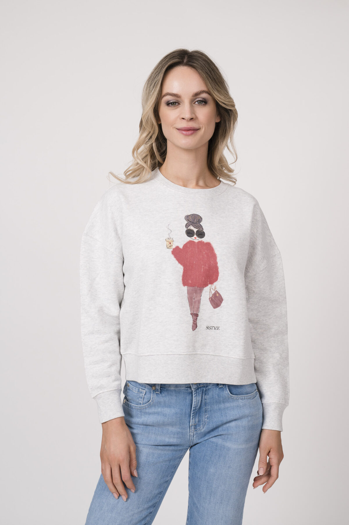 Model trägt einen Lounge-Sweater der Marke Sustyle in Hellgrau mit Motiv auf der Vorderseite. Dazu eine Jeans der Marke 7 for all mankind in Mittelblau.