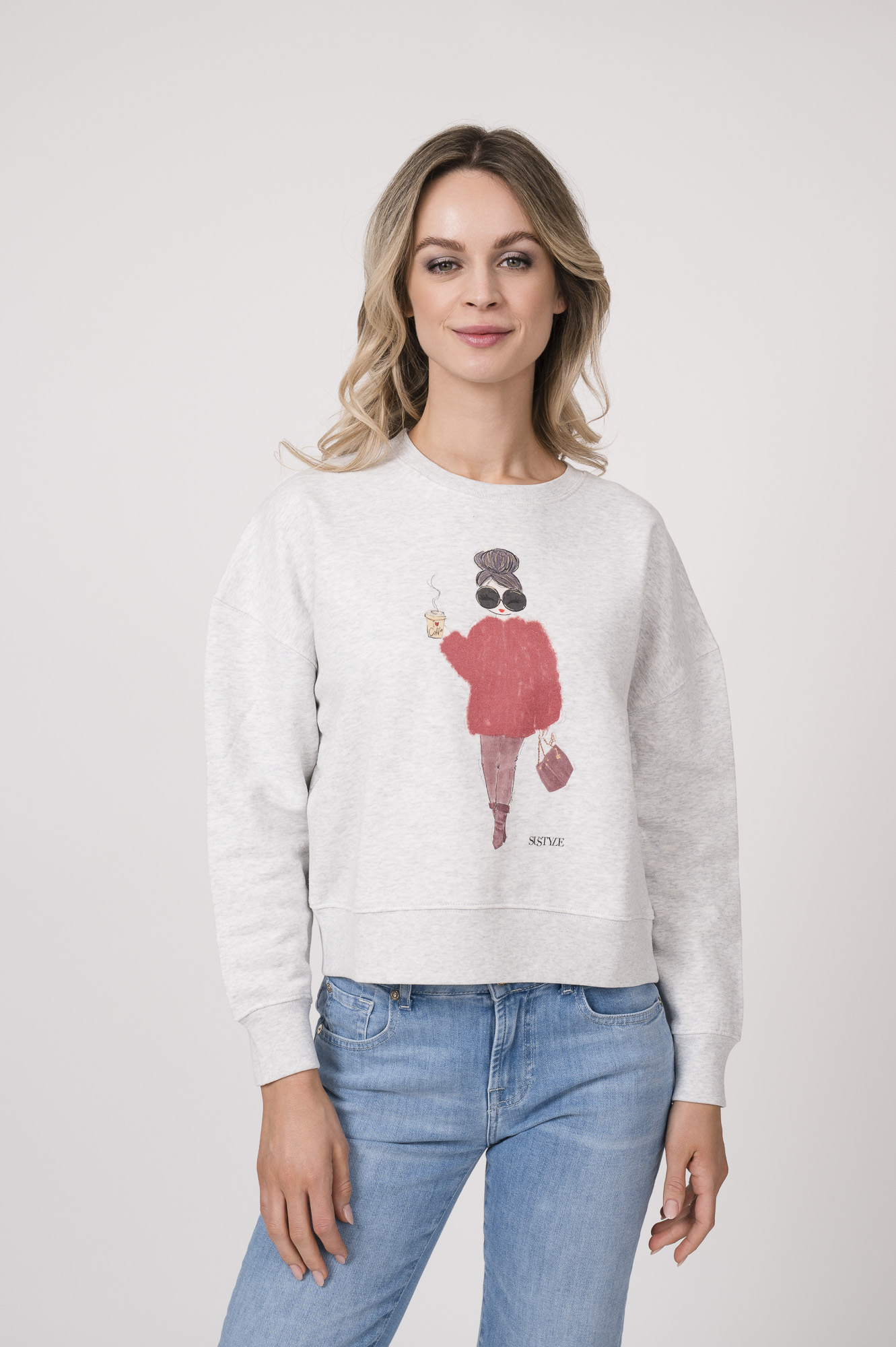 Model trägt einen Lounge-Sweater der Marke Sustyle in Hellgrau mit Motiv auf der Vorderseite. Dazu eine Jeans der Marke 7 for all mankind in Mittelblau.