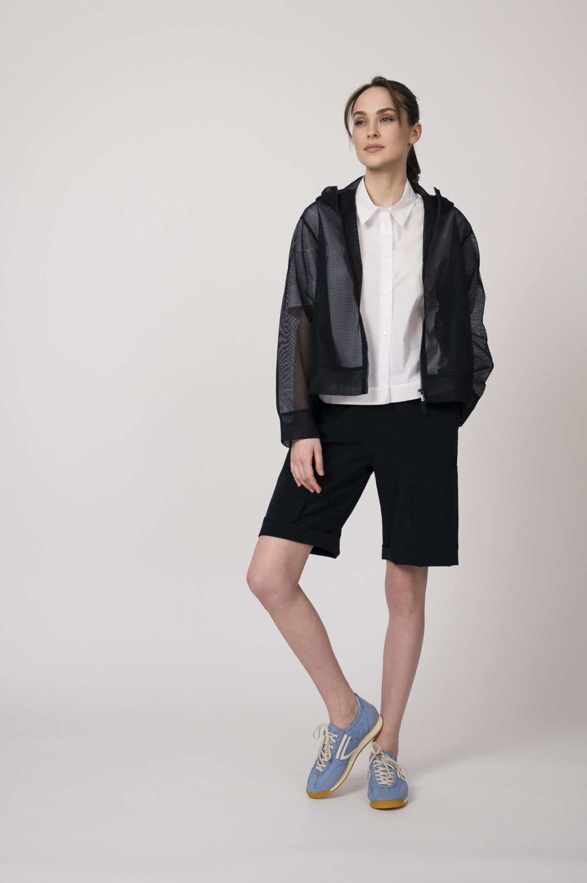 Model trägt eine marine-blaue Netzjacke mit Kapuze, eine marine-blaue Bermuda Shorts und eine weiße Kurzarmbluse der Marke Lis Lareida.