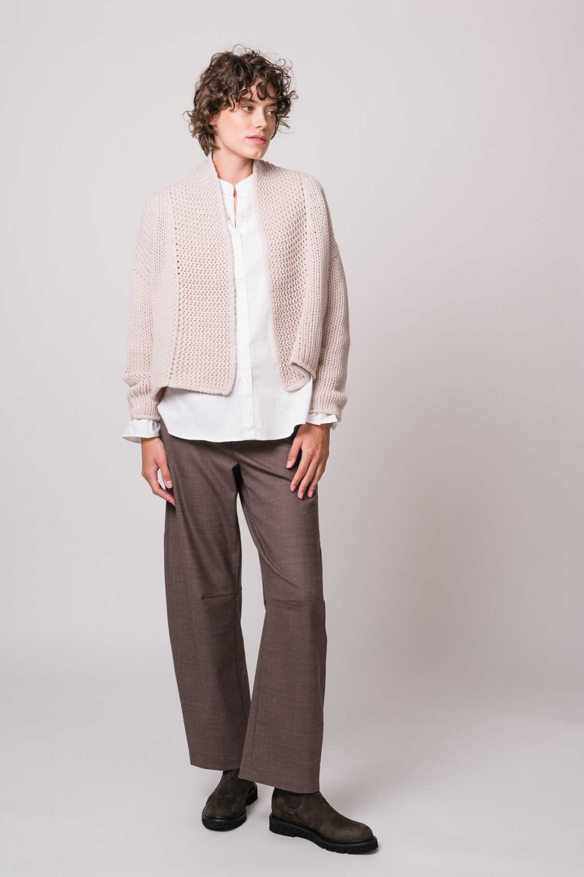 Model trägt eine weiße Bluse der Marke Rossana Diva, eine beige Strickjacke der Marke FFC und eine brauen Stoffhose der Marke Cambio.
