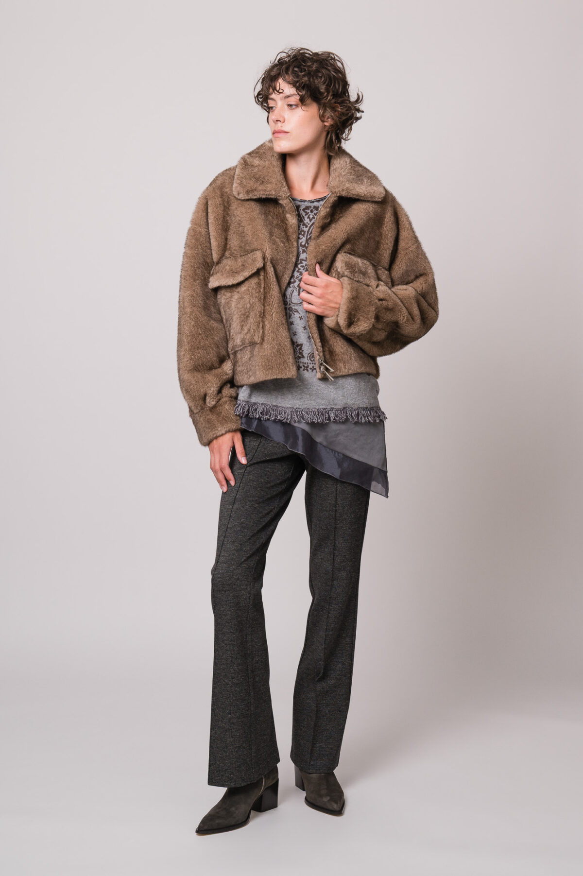 Model trägt eine braune Fake Fur Jacke der Marke Betta Corradi, ein graues Seidentop der Marke Luisa Cerano, einen grau gemusterten Pullover der Marke Grace und eine graue Stoffhose der Marke Cambio.