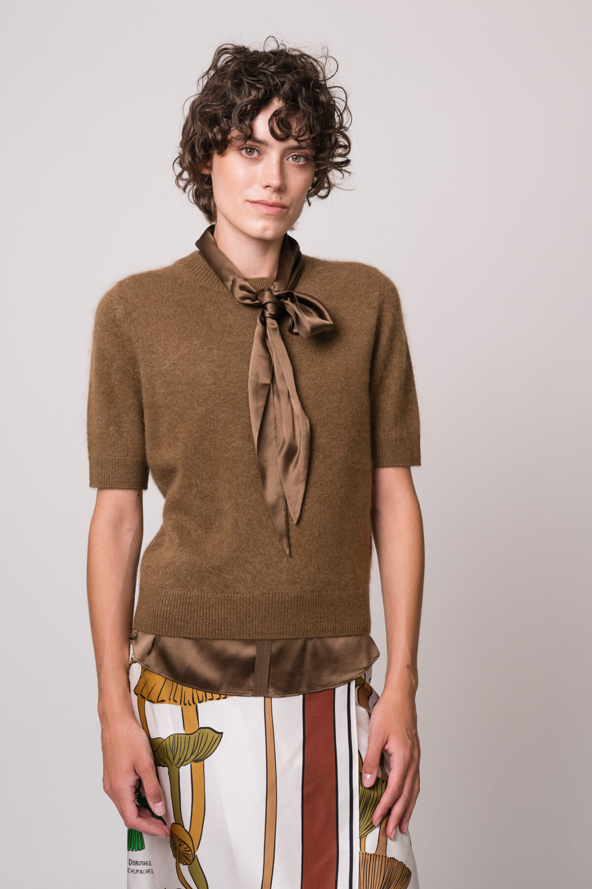 Model trägt einen braunen Kurzarmpullover, eine braune Bluse und einen gemusterten Rock der Marke Dorothee Schumacher.