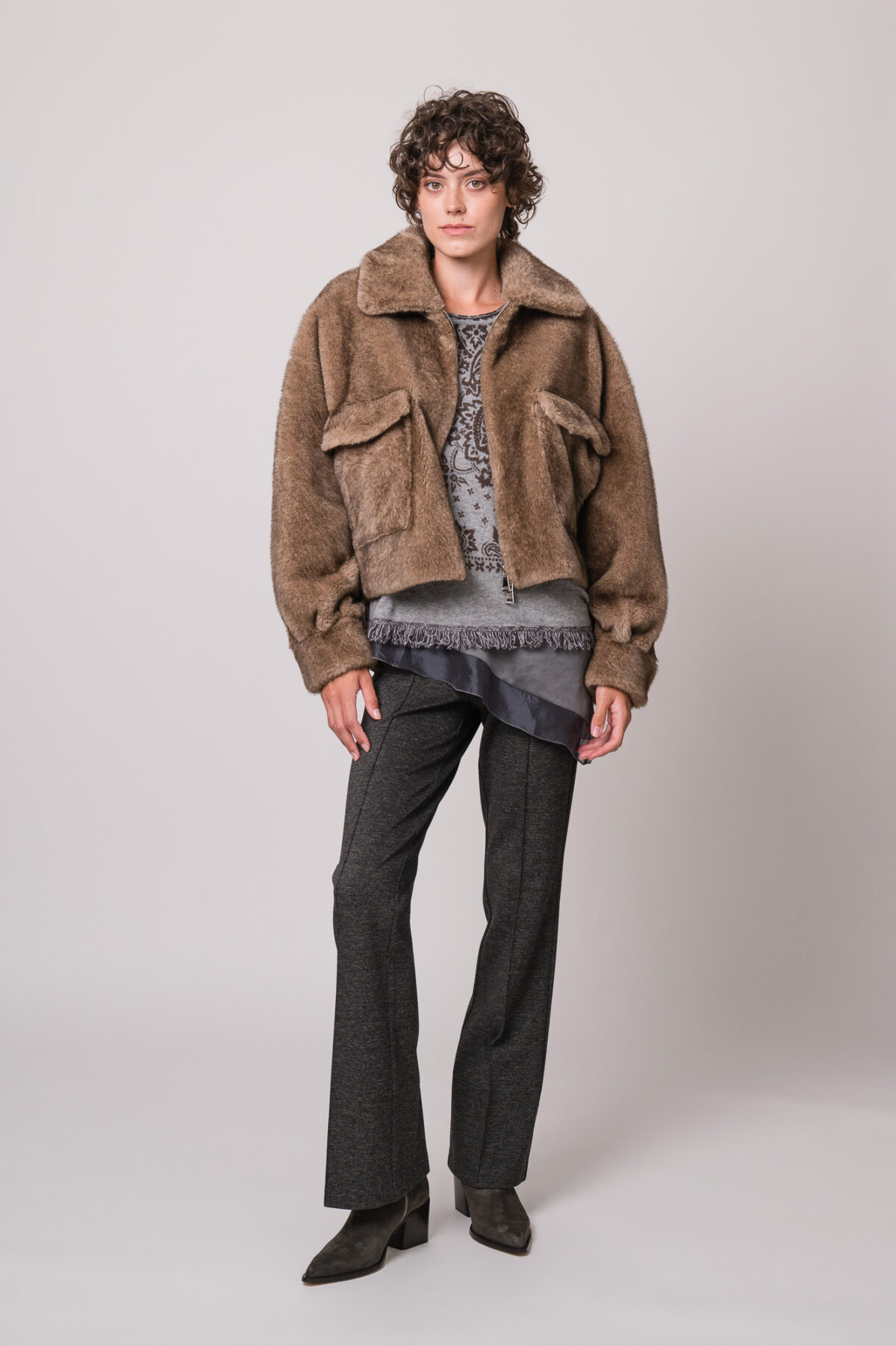 Model trägt eine braune Fake Fur Jacke der Marke Betta Corradi, ein graues Seidentop der Marke Luisa Cerano, einen grau gemusterten Pullover der Marke Grace und eine graue Stoffhose der Marke Cambio.