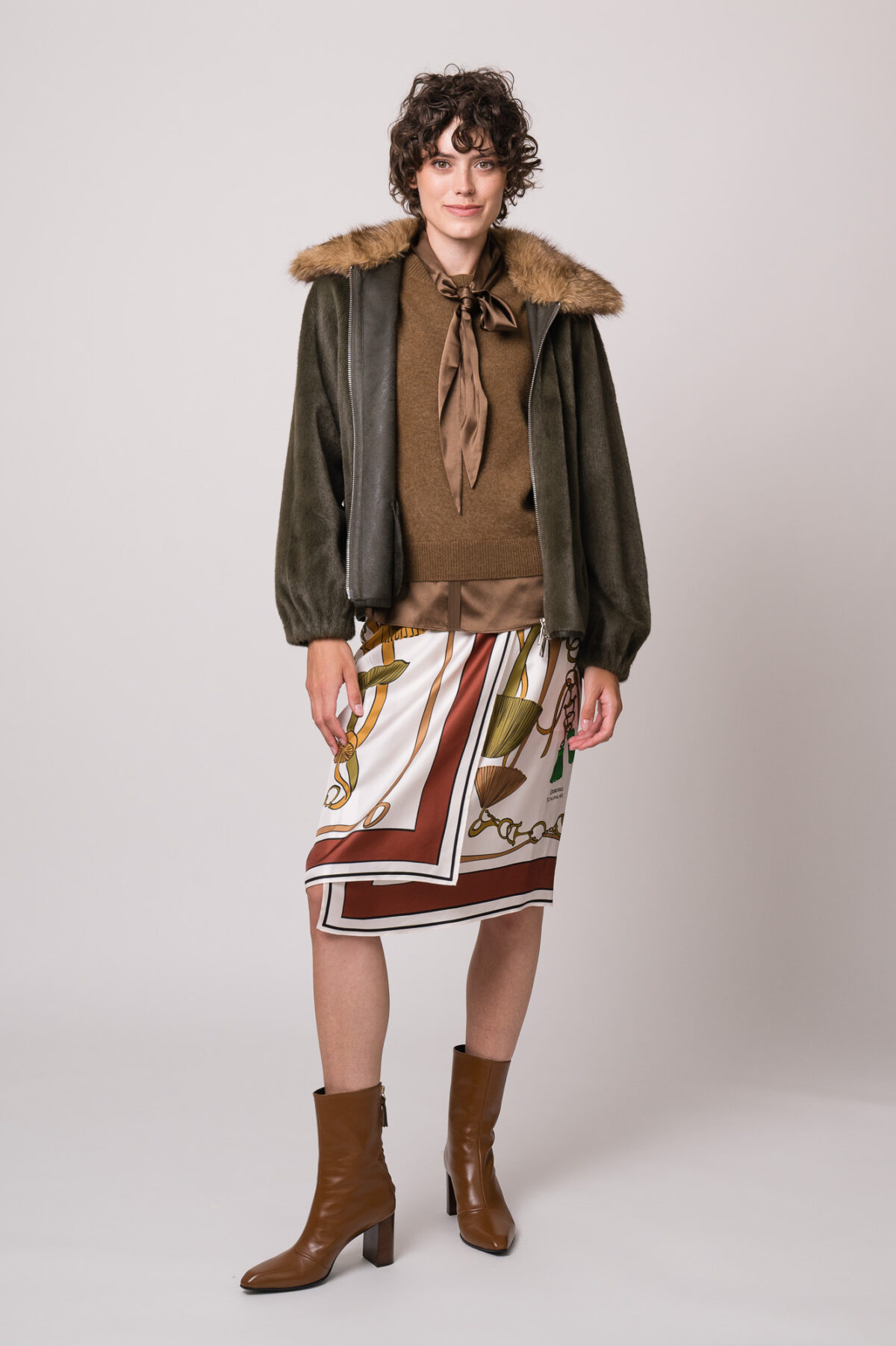 Model trägt eine grüne Jacke des Marke Betta Corradi, einen braunen Kurzarmpullover, eine braune Bluse und einen gemusterten Rock der Marke Dorothee Schumacher.