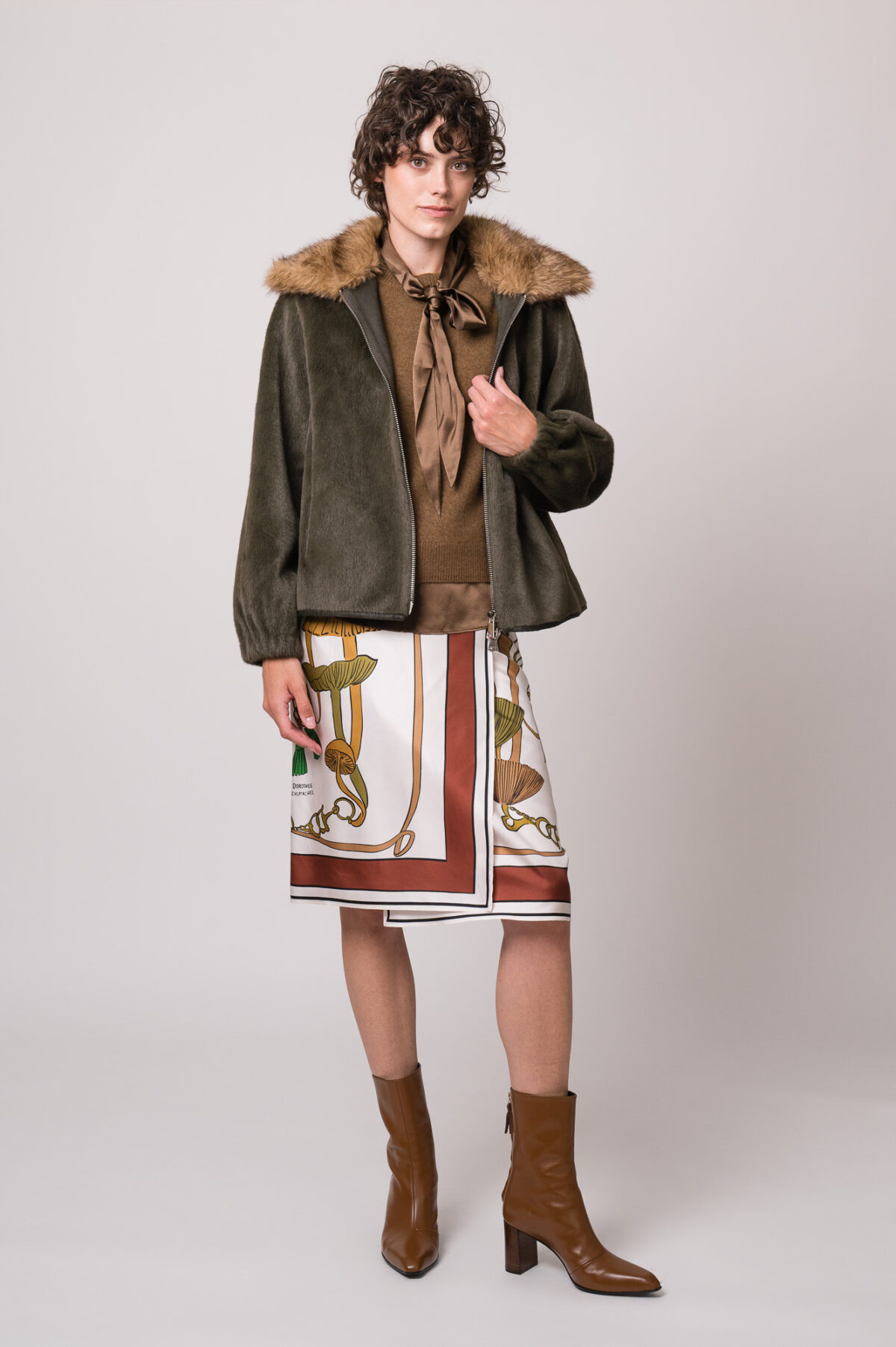 Model trägt eine grüne Jacke des Marke Betta Corradi, einen braunen Kurzarmpullover, eine braune Bluse und einen gemusterten Rock der Marke Dorothee Schumacher.