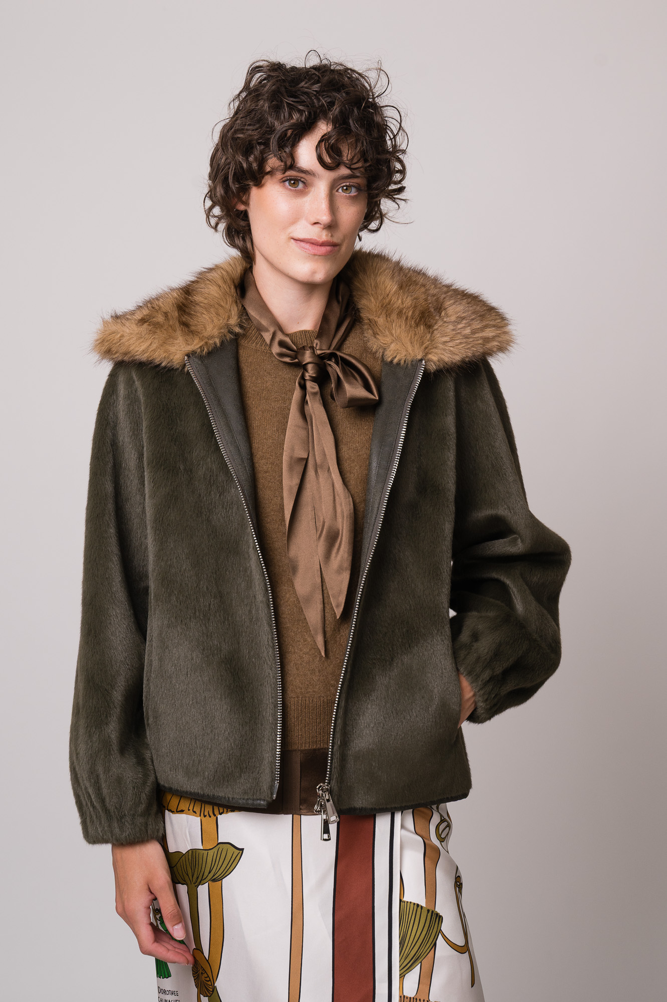 Model trägt eine grüne Jacke des Marke Betta Corradi, einen braunen Kurzarmpullover, eine braune Bluse und einen gemusterten Rock der Marke Dorothee Schumacher.