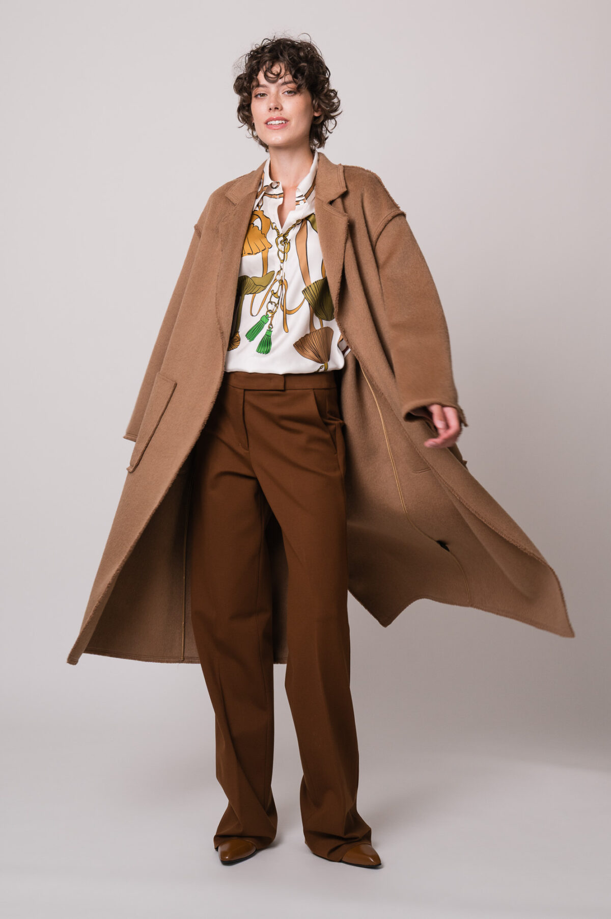 Model trägt einen braunen Mantel der Marke Lou Andrea, eine gemusterte Bluse und eine braune Hose der Marke Dorothee Schumacher.
