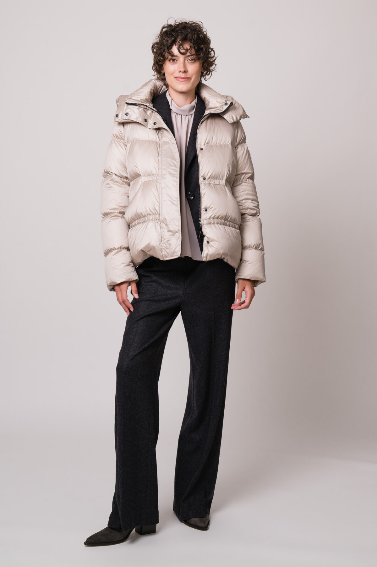 Model trägt eine beige Daunenjacke, eine helle Bluse, einen grauen Blazer und eine graue Hose der Marke Luisa Cerano.