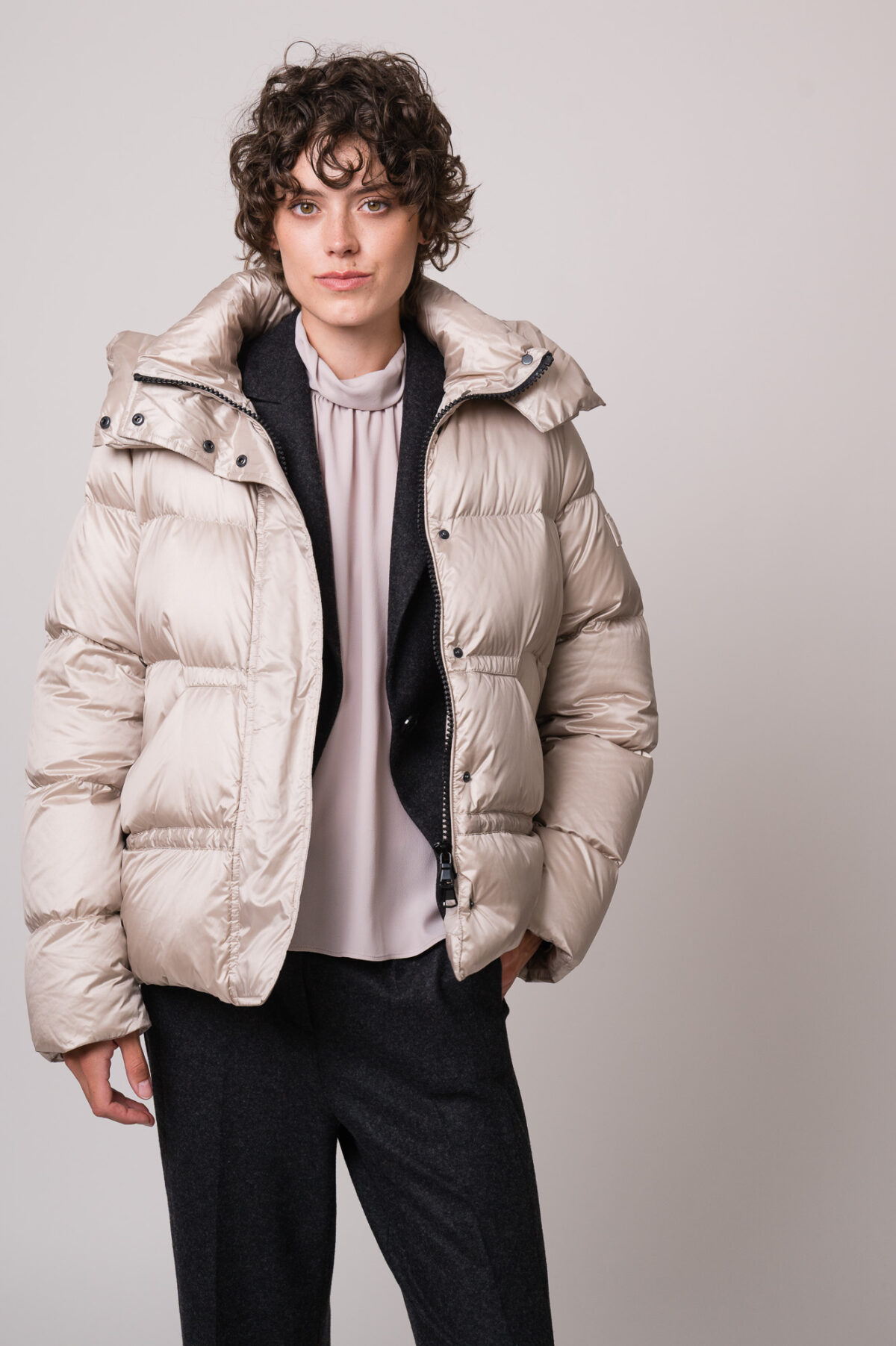 Model trägt eine beige Daunenjacke, eine helle Bluse, einen grauen Blazer und eine graue Hose der Marke Luisa Cerano.