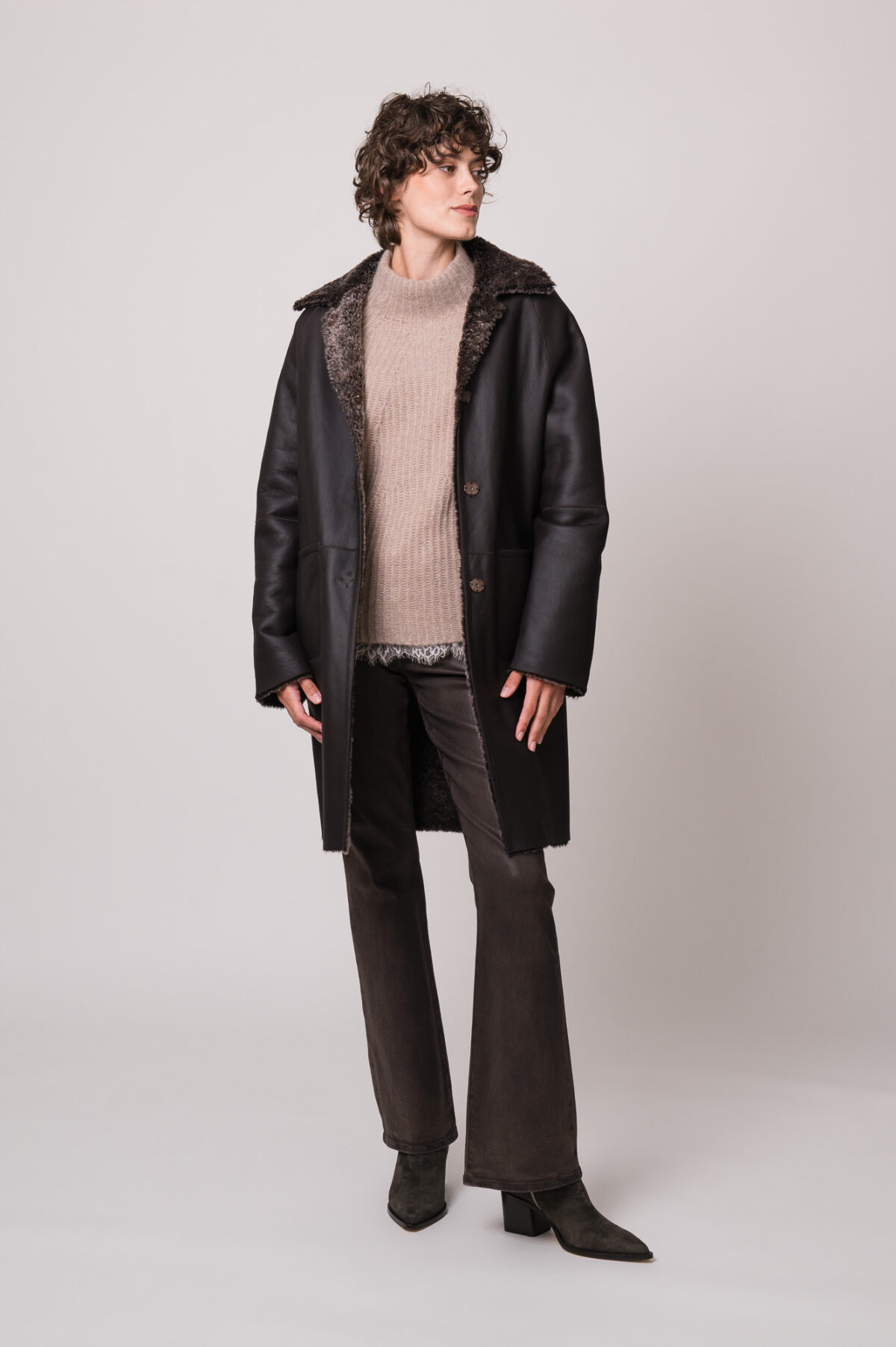 Model trägt einen braunen Lammfellmantel der Marke Hemisphere, einen beigen Pullover der Marke Hemisphere und ein beiges Top mit Spitze der Marke Allude sowie eine brauen Hose der Marke 7 for all mankind
