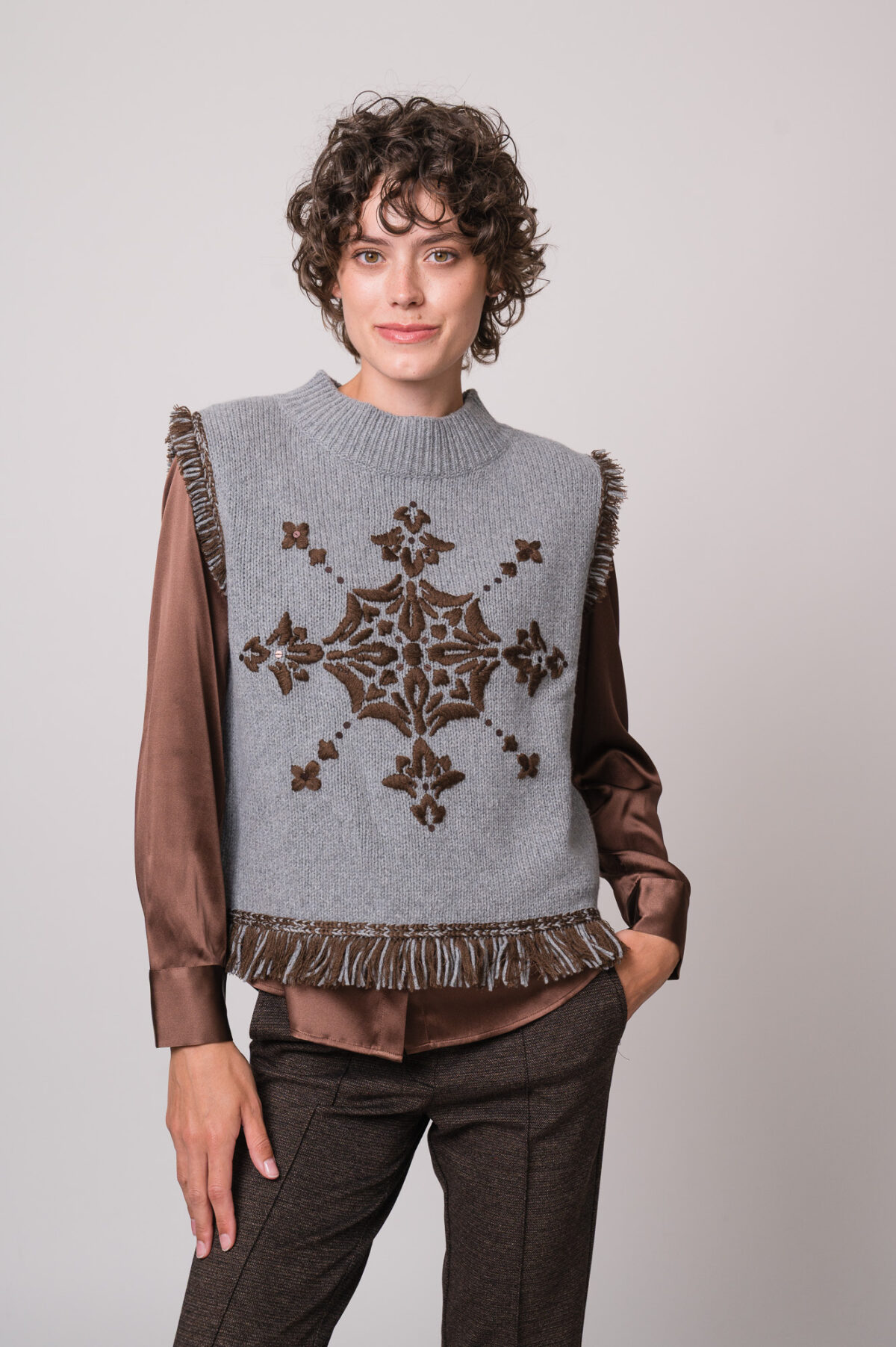 Model trägt eine braune Bluse der Marke The Mercer, einen hellgrau gemusterten Pullunder der Marke Grace.