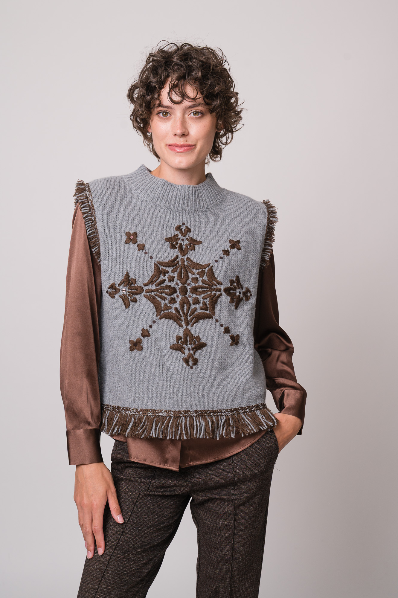 Model trägt eine braune Bluse der Marke The Mercer, einen hellgrau gemusterten Pullunder der Marke Grace.