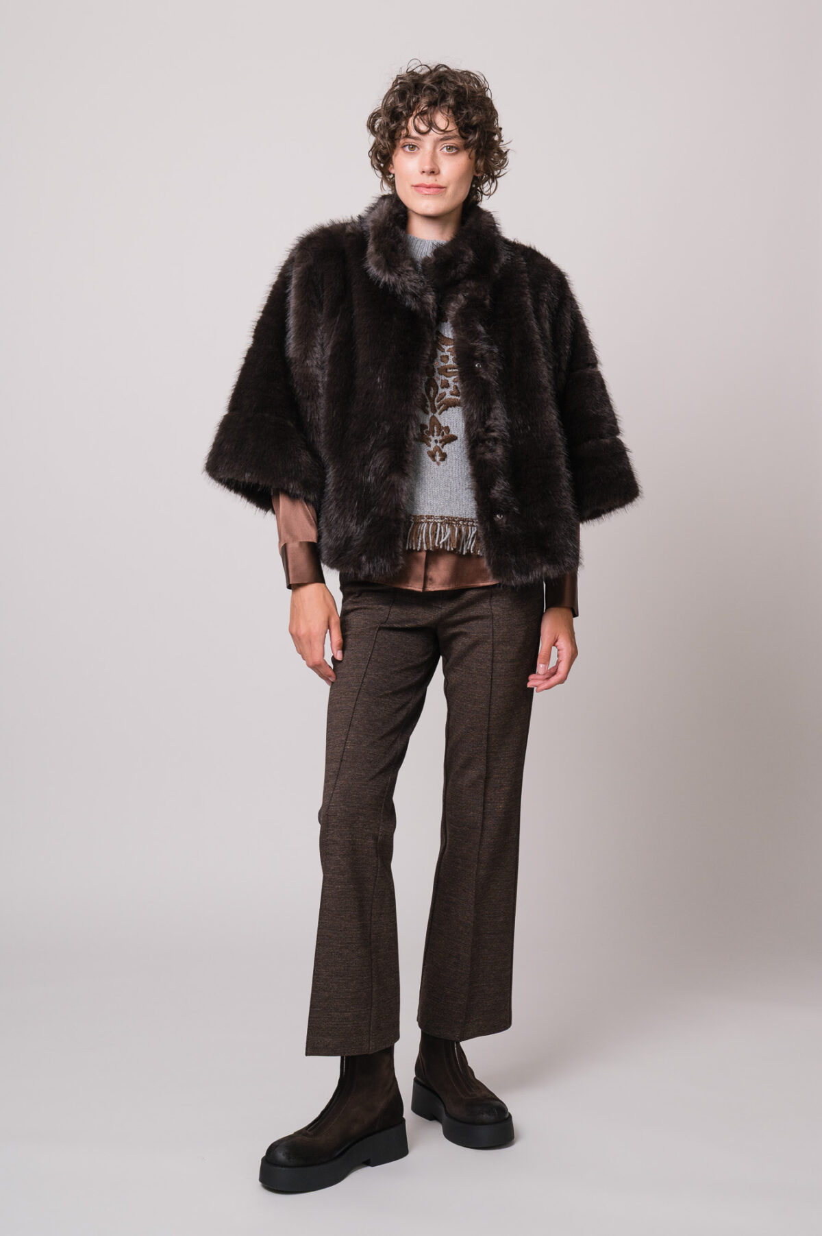 Model trägt eine braune Bluse der Marke The Mercer, einen hellgrau gemusterten Pullunder der Marke Grace sowie eine dunkelbraune Fake Für Jacke der Marke Luisa Cerano und eine braune Hose der Marke Cambio.
