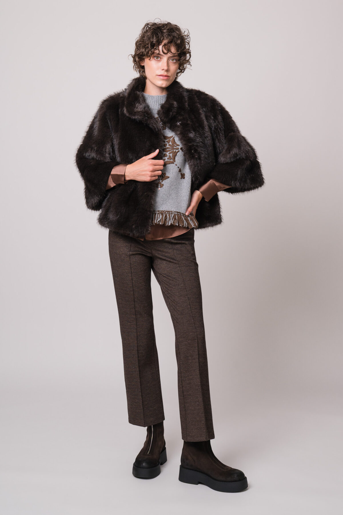 Model trägt eine braune Bluse der Marke The Mercer, einen hellgrau gemusterten Pullunder der Marke Grace sowie eine dunkelbraune Fake Für Jacke der Marke Luisa Cerano und eine braune Hose der Marke Cambio.