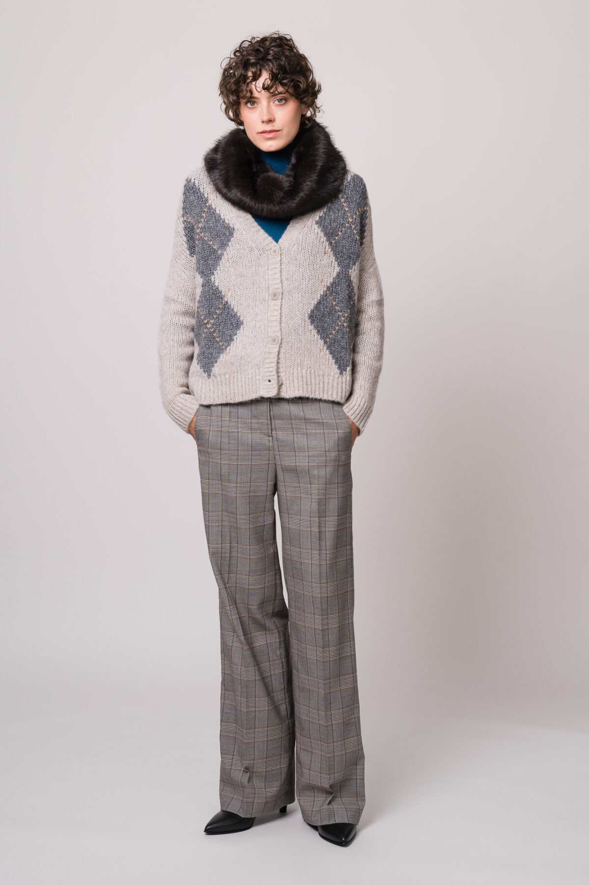 Model trägt eine grau gemusterte Weste und einen türkisfarbigen Rollkragenpullover sowie einen Fake Fur Schlauchschal und eine grau karierte Stoffhose der Marke Luisa Cerano.
