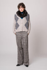 Model trägt eine grau gemusterte Weste und einen türkisfarbigen Rollkragenpullover sowie einen Fake Fur Schlauchschal und eine grau karierte Stoffhose der Marke Luisa Cerano.