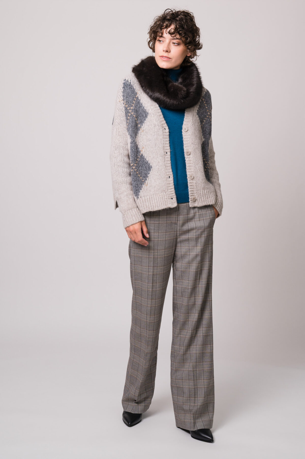 Model trägt eine grau gemusterte Weste und einen türkisfarbigen Rollkragenpullover sowie einen Fake Fur Schlauchschal und eine grau karierte Stoffhose der Marke Luisa Cerano.