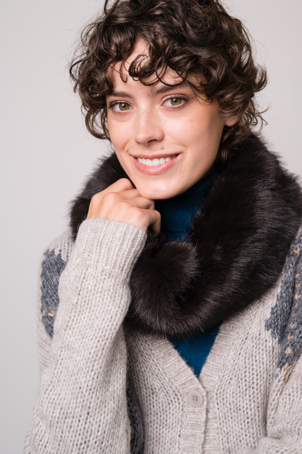 Model trägt eine grau gemusterte Weste und einen türkisfarbigen Rollkragenpullover sowie einen Fake Fur Schlauchschal der Marke Luisa Cerano.