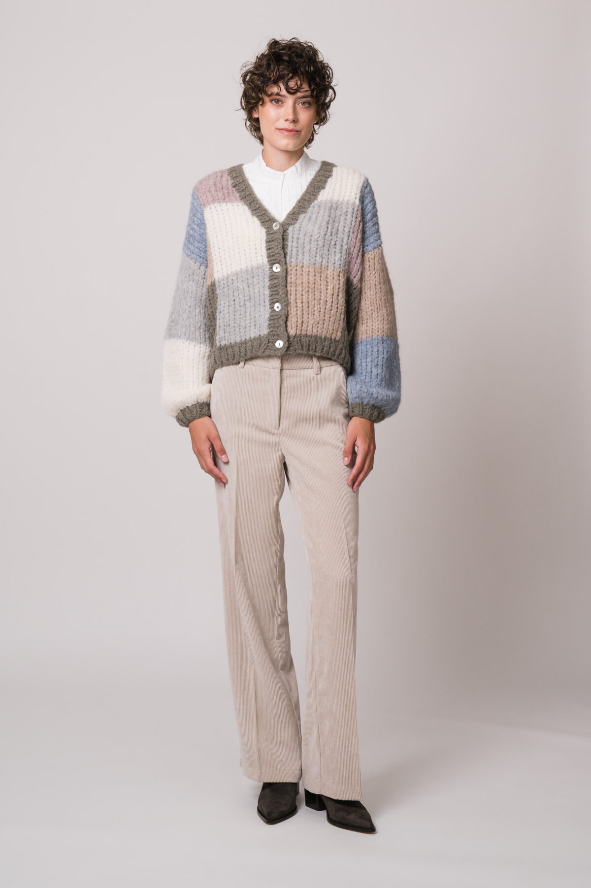 Model trägt eine bunte Strickweste der Marke Michele & Hoven, eine weiße Bluse der Marke SOSUE und eine beige Kordhose der Marke Cambio.