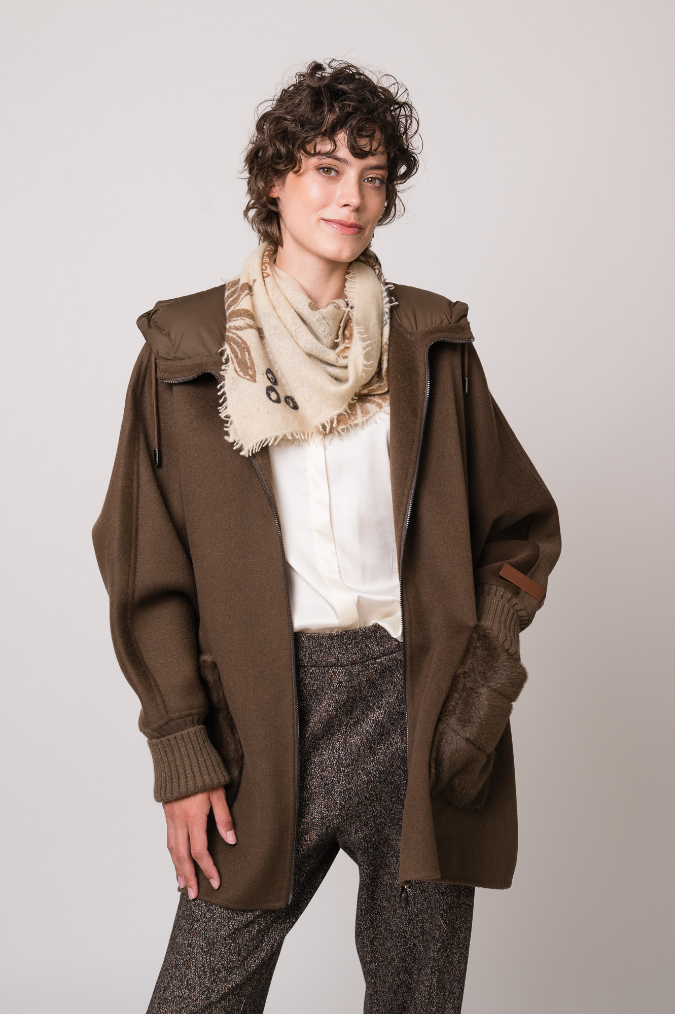 Model trägt eine braune Jacke der Marke Lou Andrea, eine natur-farbige Bluse der Marke The Mercer, eine dunkelbraune Hose der Marke Cambio und einen beigen Schal der Marke Mouleta.