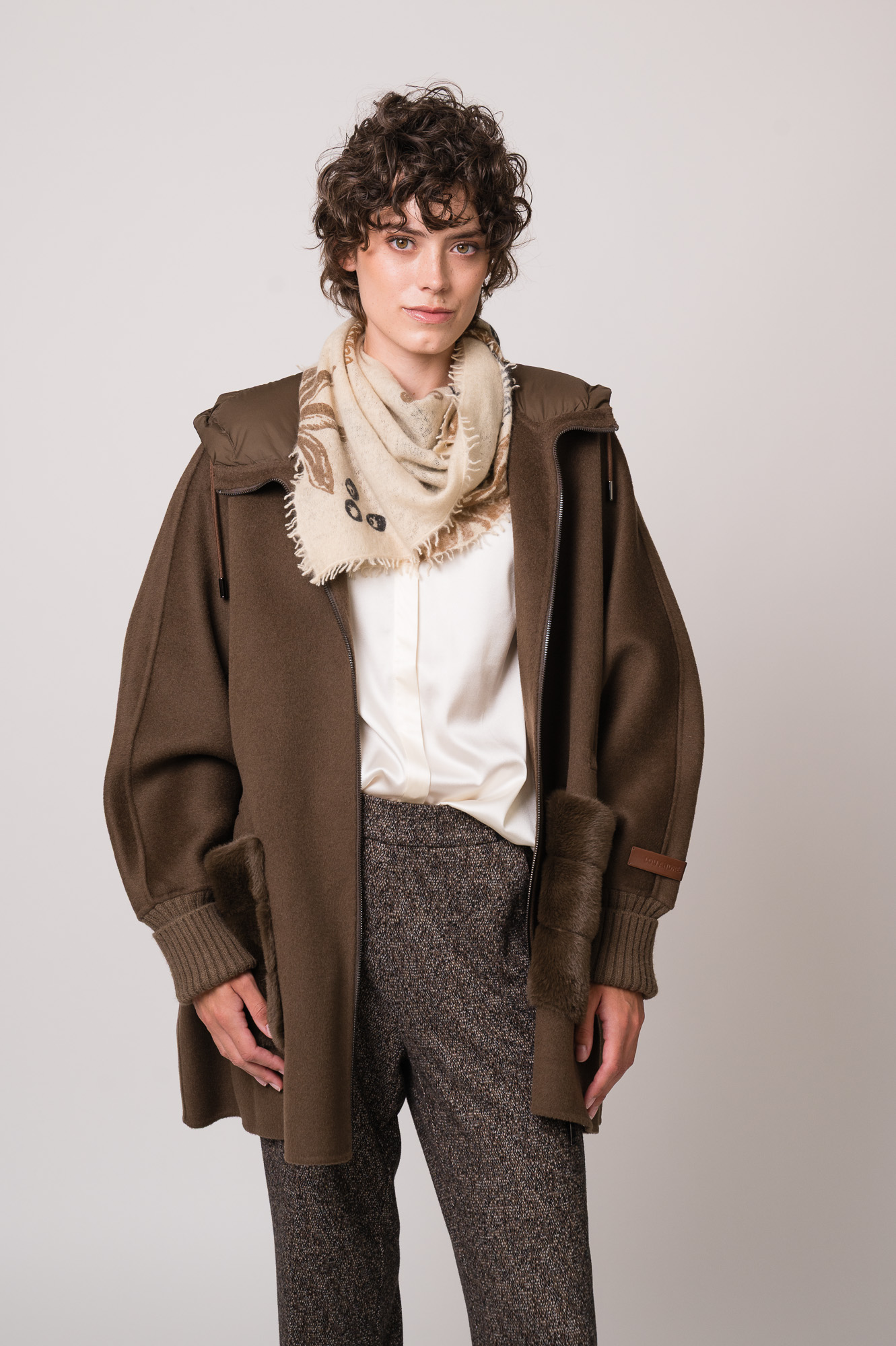 Model trägt eine braune Jacke der Marke Lou Andrea, eine natur-farbige Bluse der Marke The Mercer, eine dunkelbraune Hose der Marke Cambio und einen beigen Schal der Marke Mouleta.