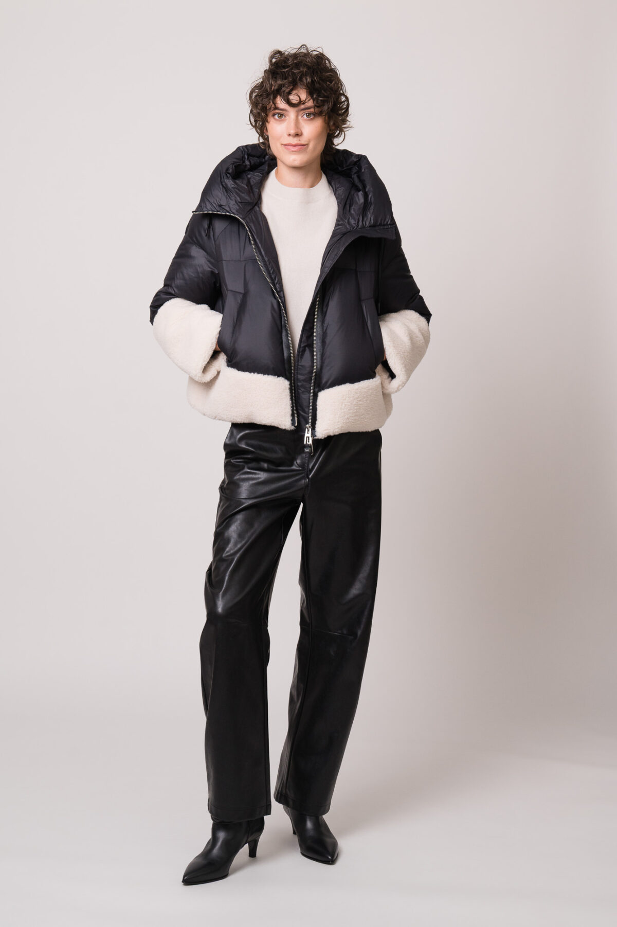 Model trägt eine schwarze Daunenjacke mit weißem Fellbesatz der Marke Duno, einen beigen Pullover der Marke Herzens und eine schwarze Hose der Marke Cambio.