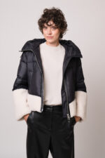 Model trägt eine schwarze Daunenjacke mit weißem Fellbesatz der Marke Duno, einen beigen Pullover der Marke Herzens und eine schwarze Hose der Marke Cambio.