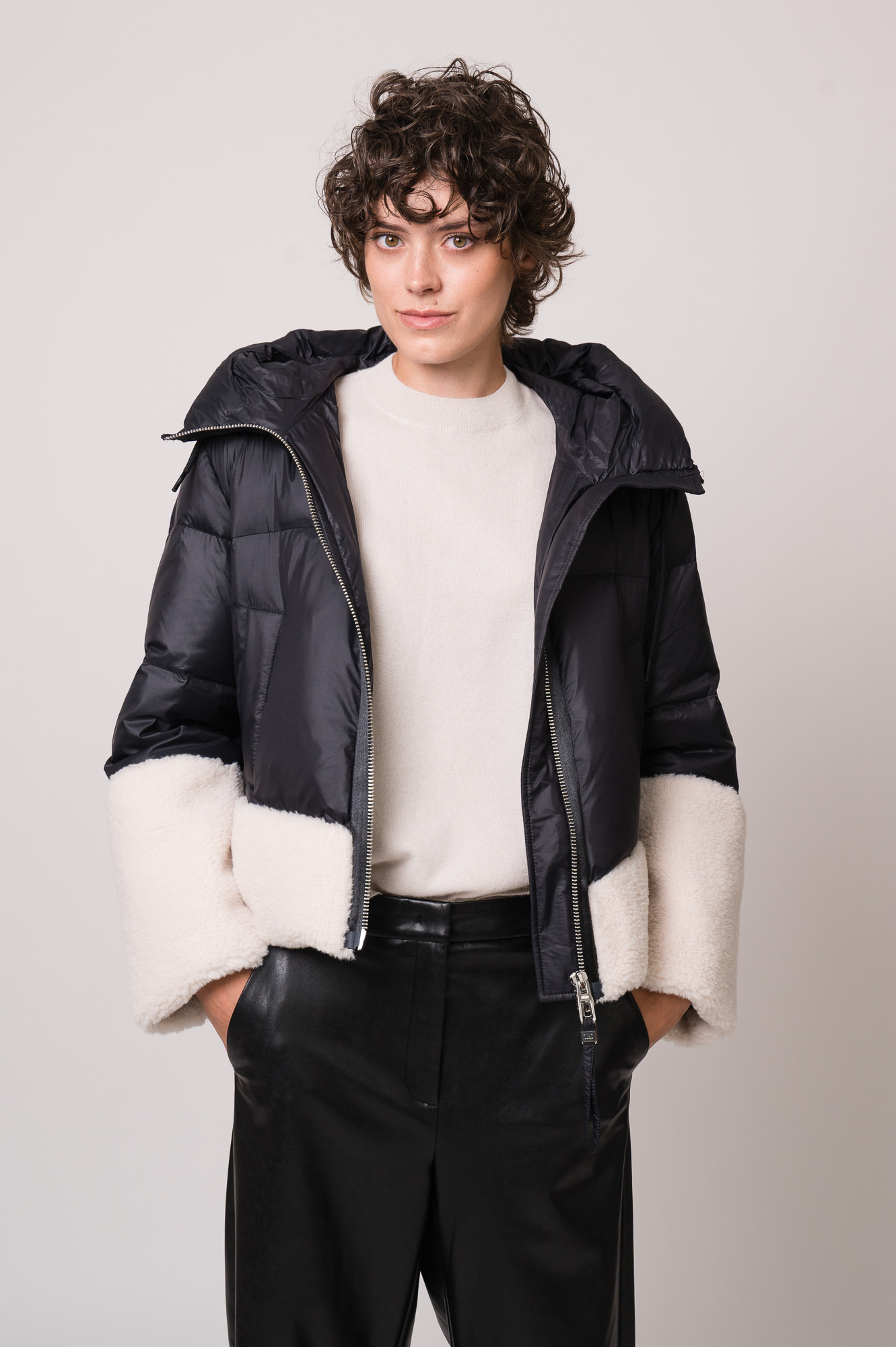 Model trägt eine schwarze Daunenjacke mit weißem Fellbesatz der Marke Duno, einen beigen Pullover der Marke Herzens und eine schwarze Hose der Marke Cambio.